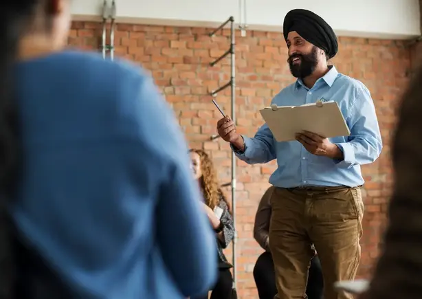 A man holding a clipboard and pen speaks to a seated group in a room with brick walls.